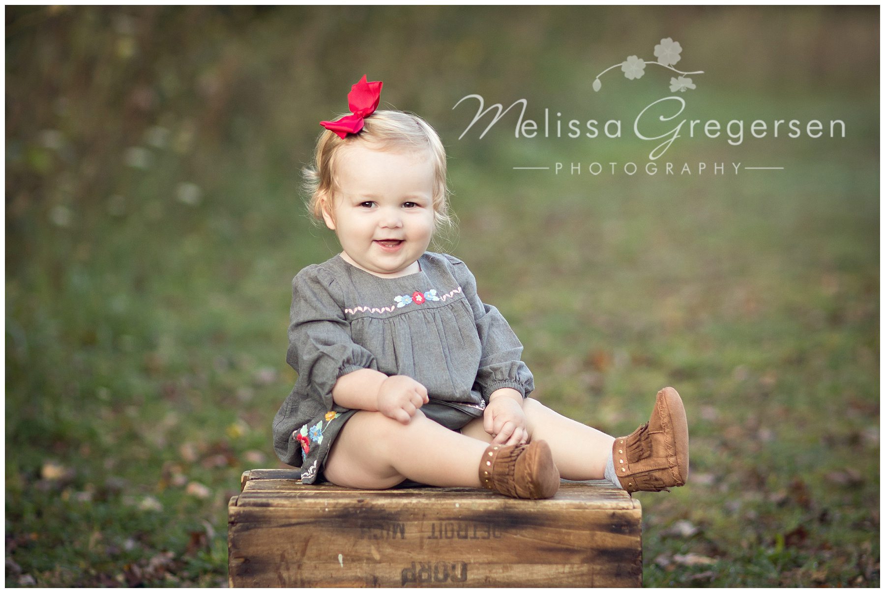 Adorable baby sitting on a brown box for photography session with family wearing a red bow