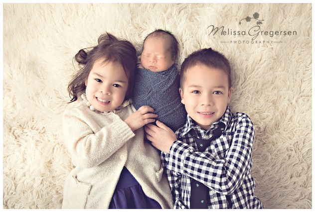 Newborn with both older siblings on simple off white rug.